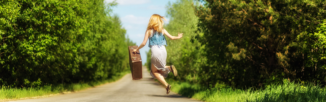 An image of a girl jumping in the middle of country road with a suitcase